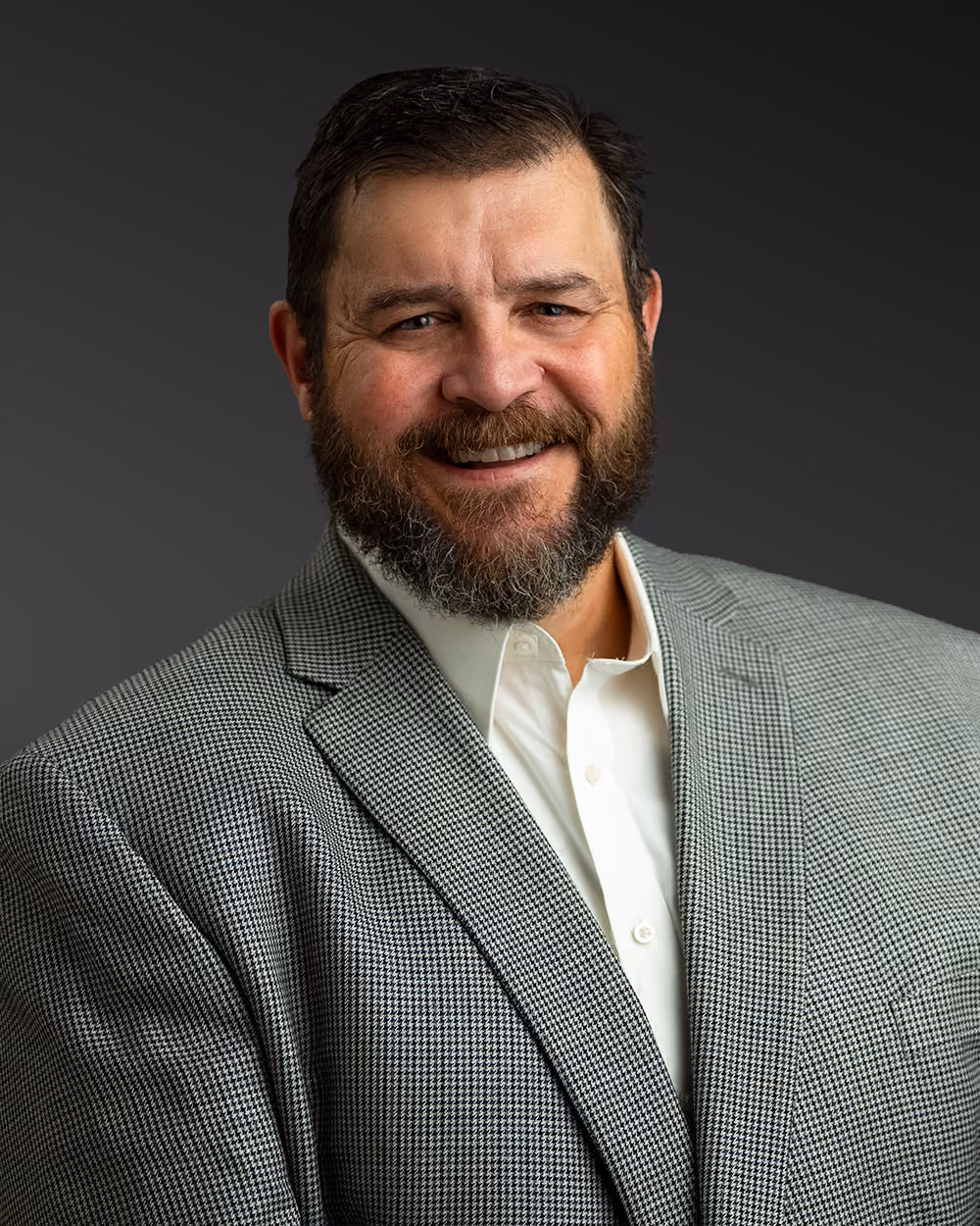 Smiling bearded man in a gray houndstooth blazer and white shirt against dark background.