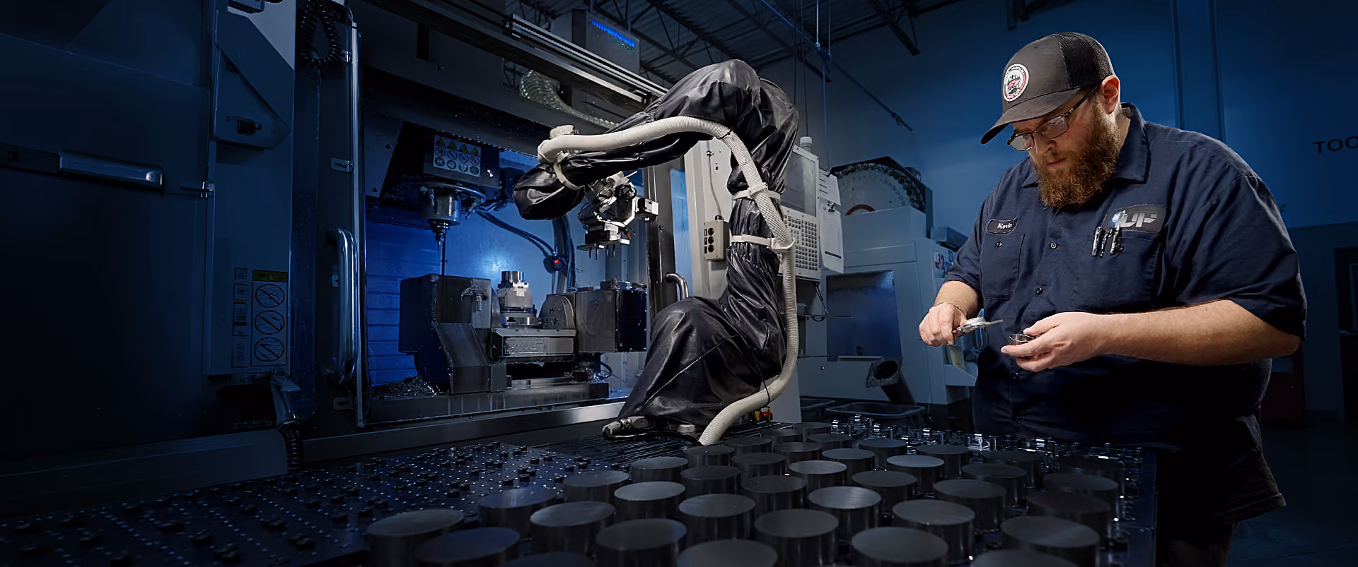 Factory worker inspecting a metal part beside a CNC machine and a robotic arm in a manufacturing setting.