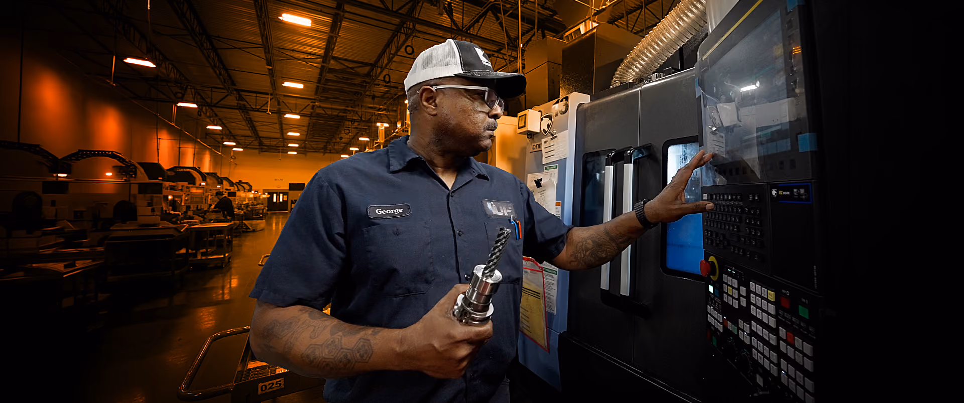 Factory worker in uniform and cap operating a machine with a control panel in an industrial setting.