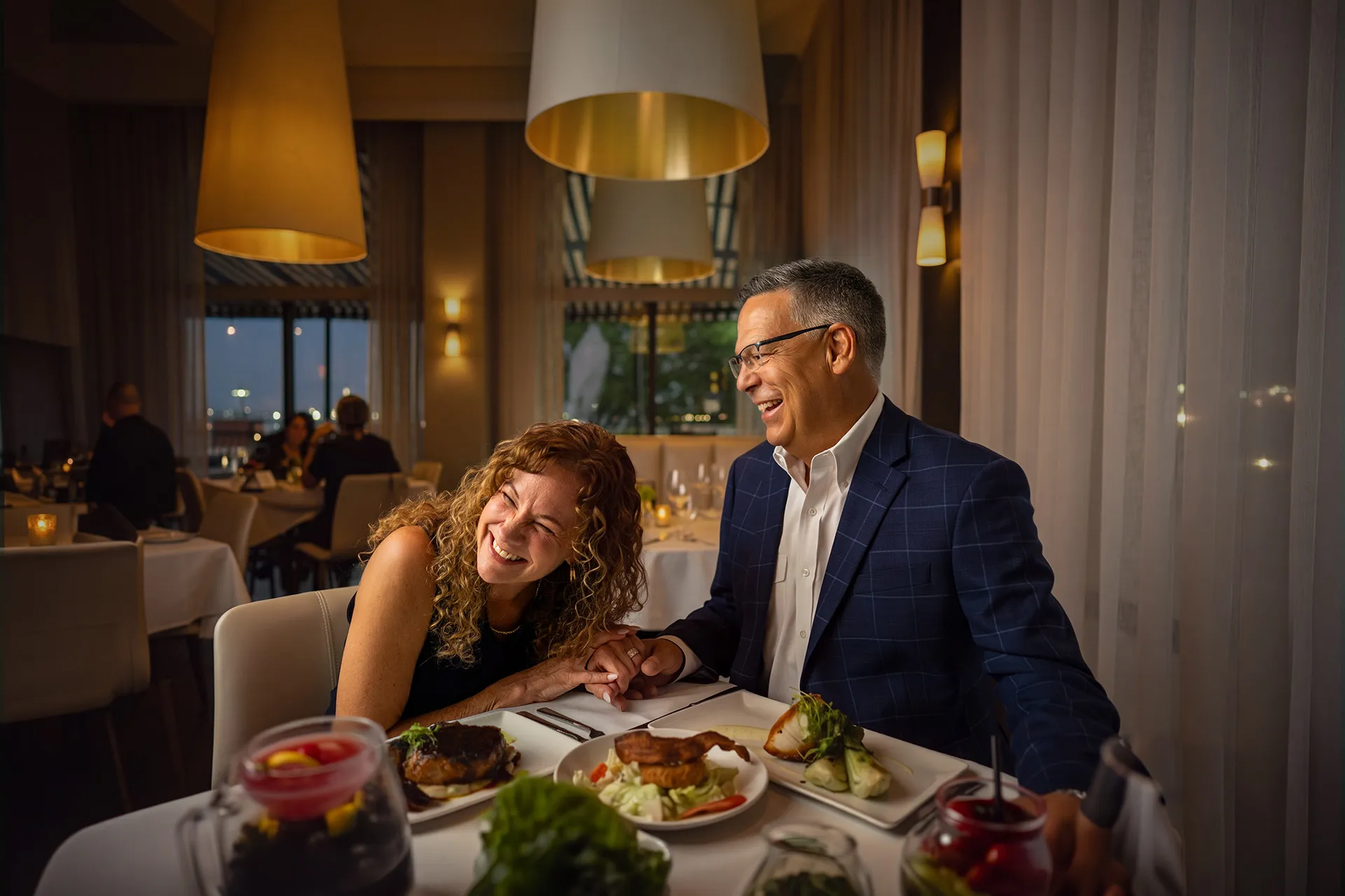 A smiling middle-aged couple holding hands and laughing while dining at a restaurant, with plates of food in front of them and elegant indoor lighting.