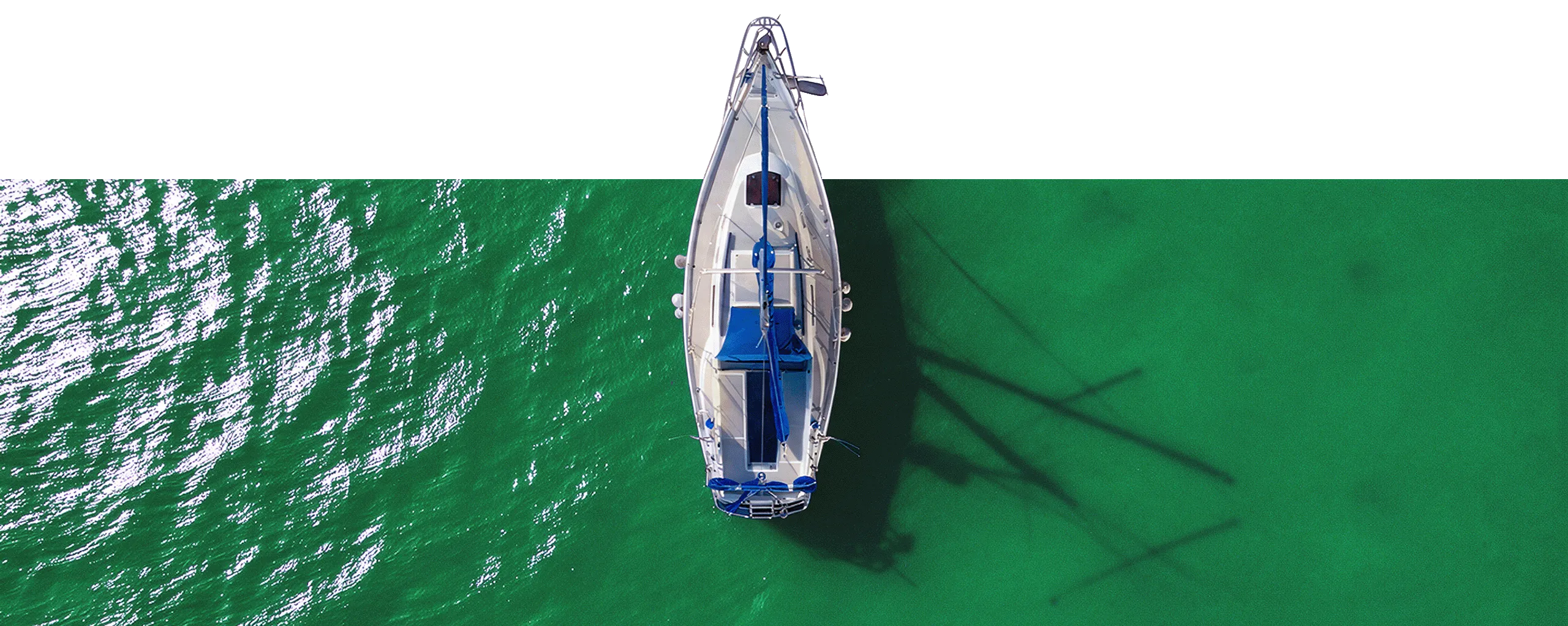 A white sailboat with blue accents seen from directly above floating on green water with sunlight reflections and the boat's shadow visible below.