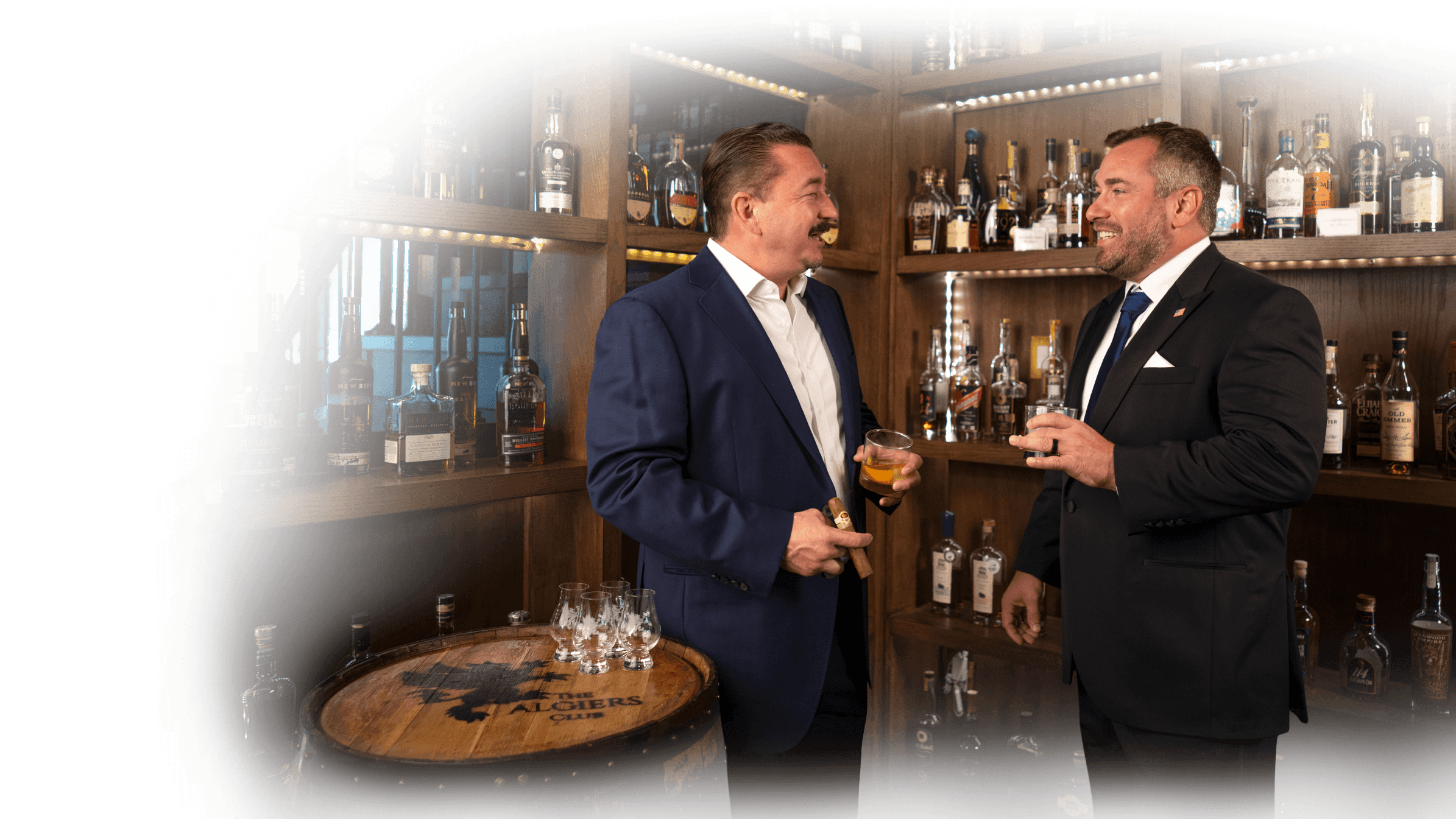 Two men in suits enjoying drinks and a cigar while conversing in a cozy, wood-paneled bar with shelves of liquor bottles and a wooden barrel with glasses on top labeled The Algiers Club.
