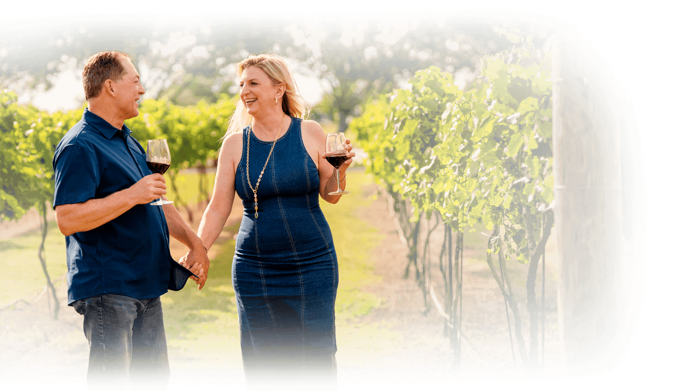Couple holding wine glasses walking through a vineyard