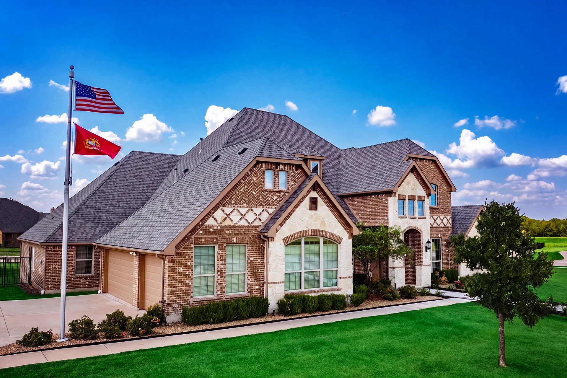 Large two-story brick house with light stone accents, a three-car garage, manicured bushes, a green lawn, and two flagpoles displaying the American and Marine Corps flags under a bright blue sky with scattered clouds.