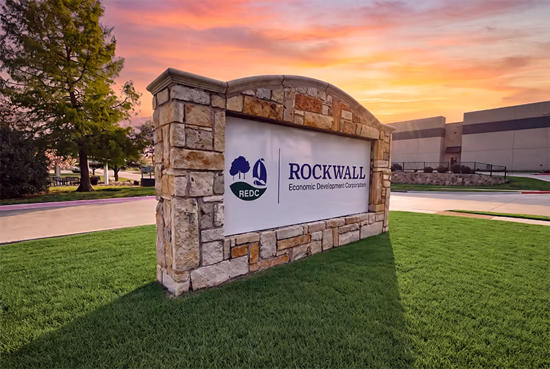 Stone sign for Rockwall Economic Development Corporation on green lawn at sunset with a tree and industrial buildings in the background.