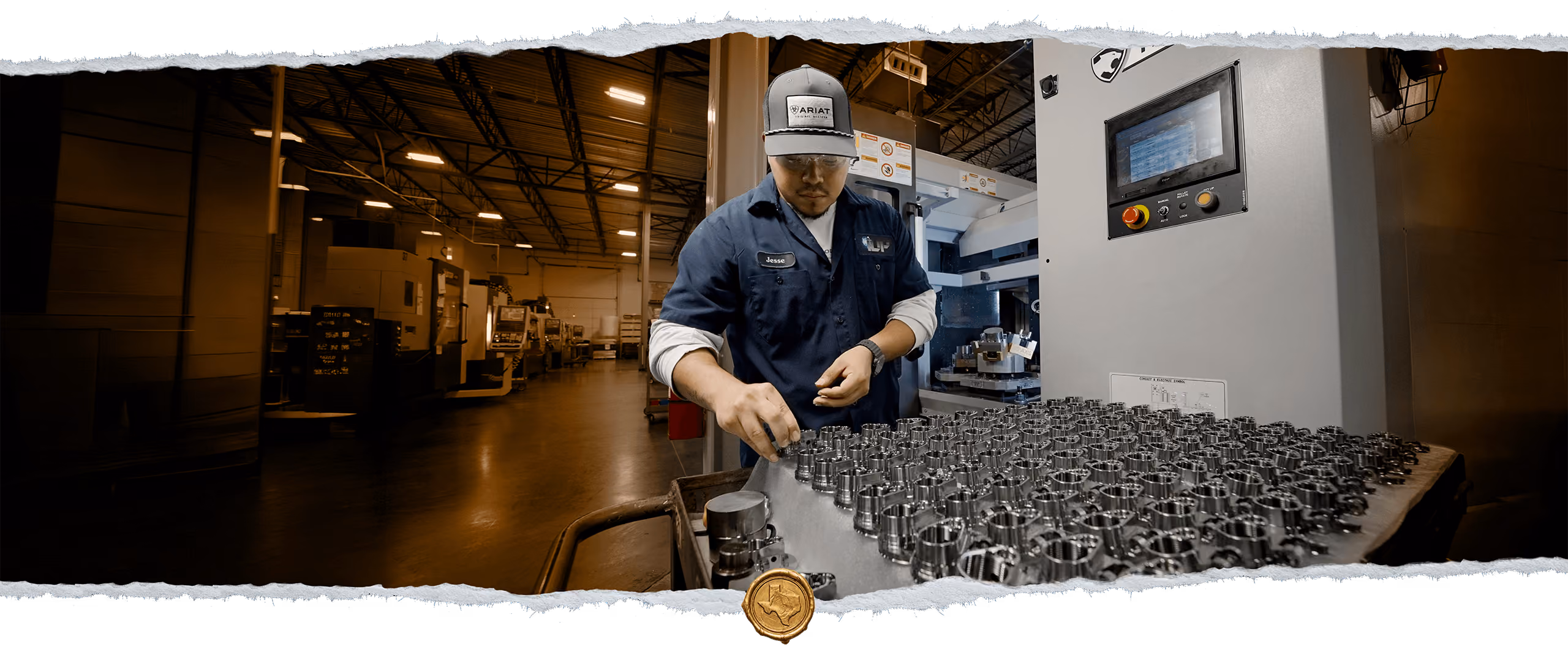 A factory worker wearing a navy shirt and a cap carefully arranges metal components on a tray in a manufacturing facility with machinery and industrial equipment in the background.