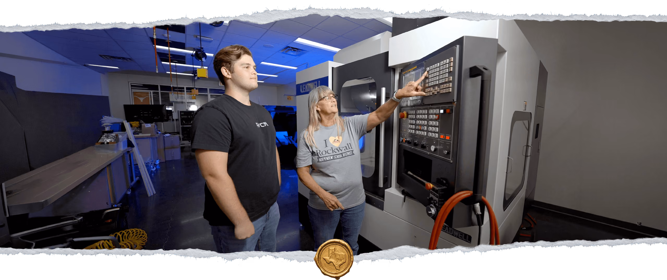 A woman wearing an I ♥ Rockwall t-shirt is teaching a young man how to operate a large industrial machine with a control panel in a workshop.