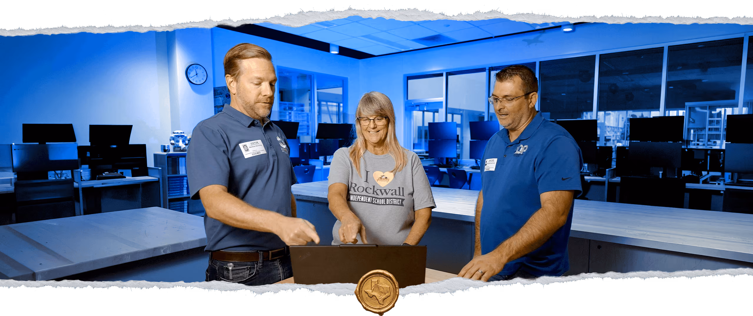 Three people standing around a laptop in a modern office or workspace; two men wear blue polo shirts with name tags, and a woman in the middle wears a gray 'I love Rockwall Independent School District' T-shirt, all focused on the laptop screen.