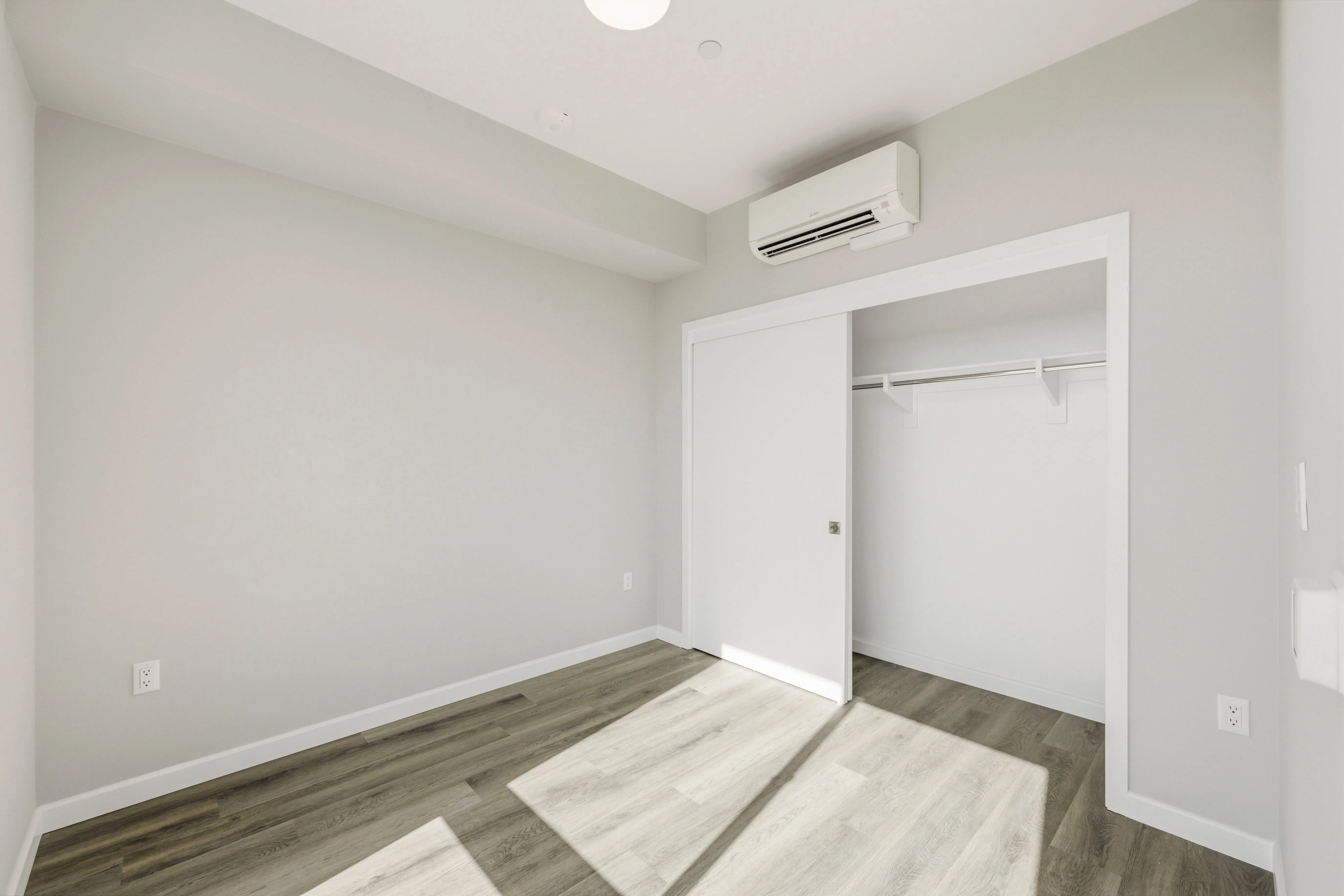 Empty room with gray walls, wood flooring, sliding closet doors, and a wall-mounted air conditioner.