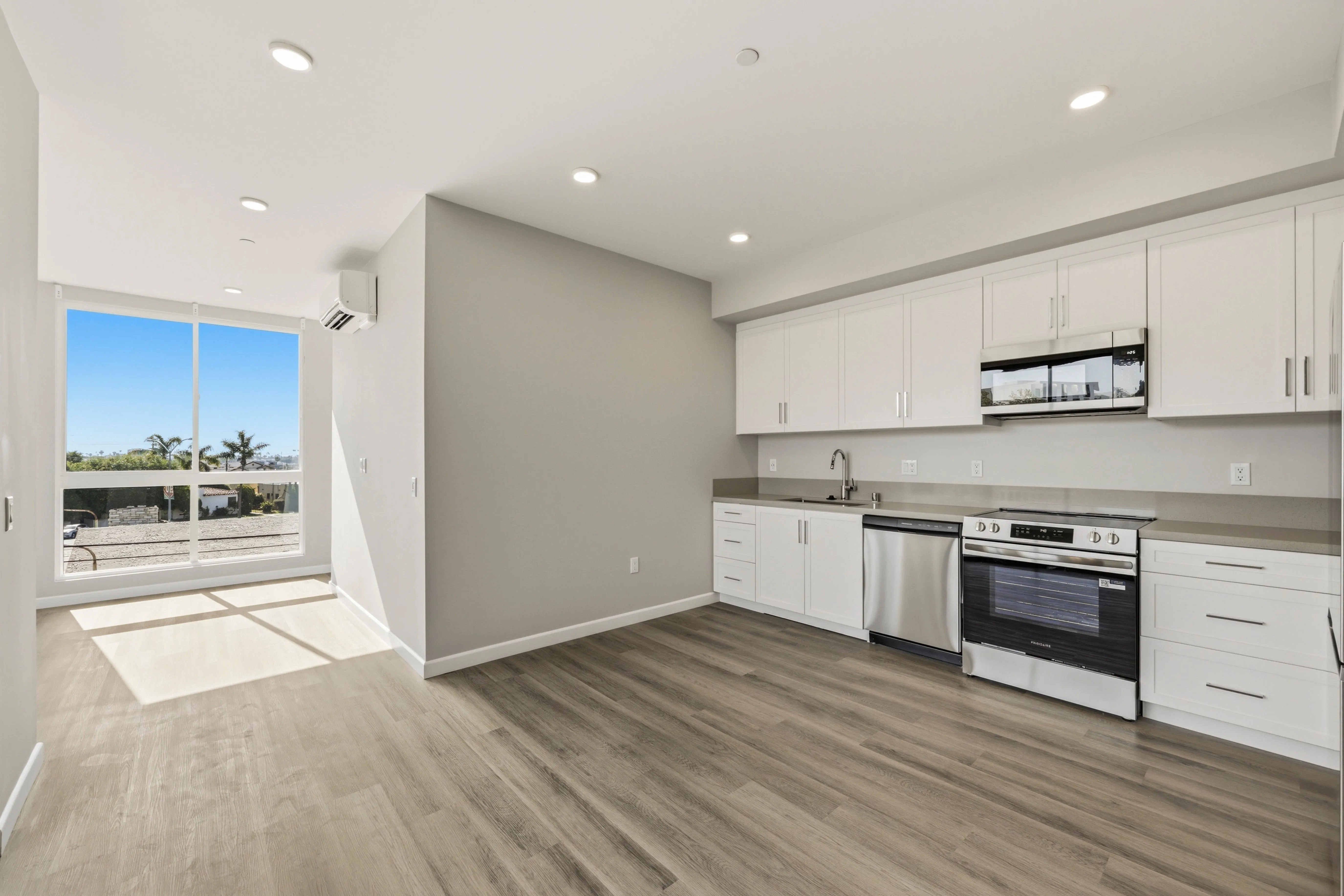 Modern kitchen with white cabinets, stainless steel appliances, wood floor, and large window with sunlight.