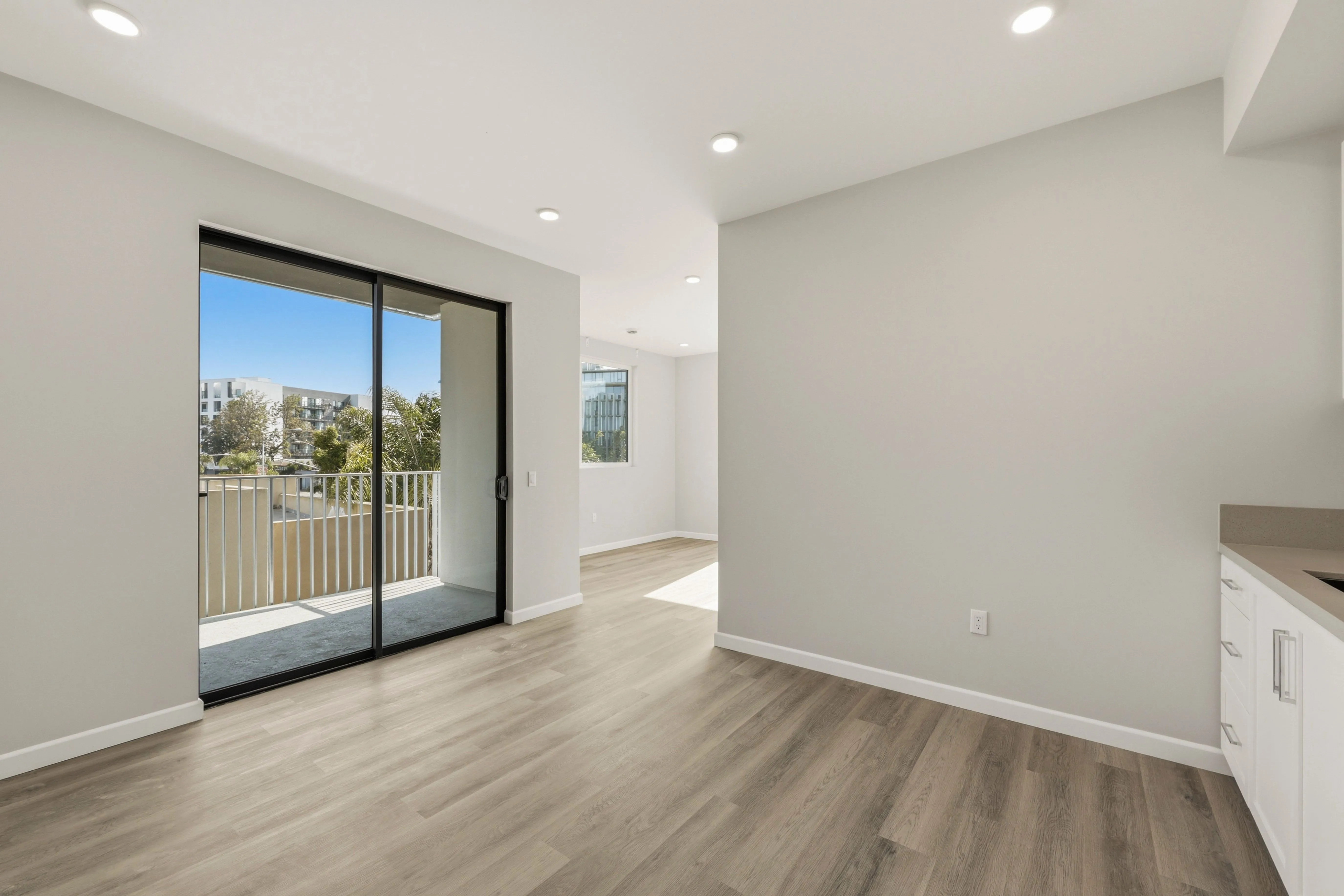 Empty modern apartment with light wood floors, white walls, recessed lighting, and a sliding glass door to a balcony.