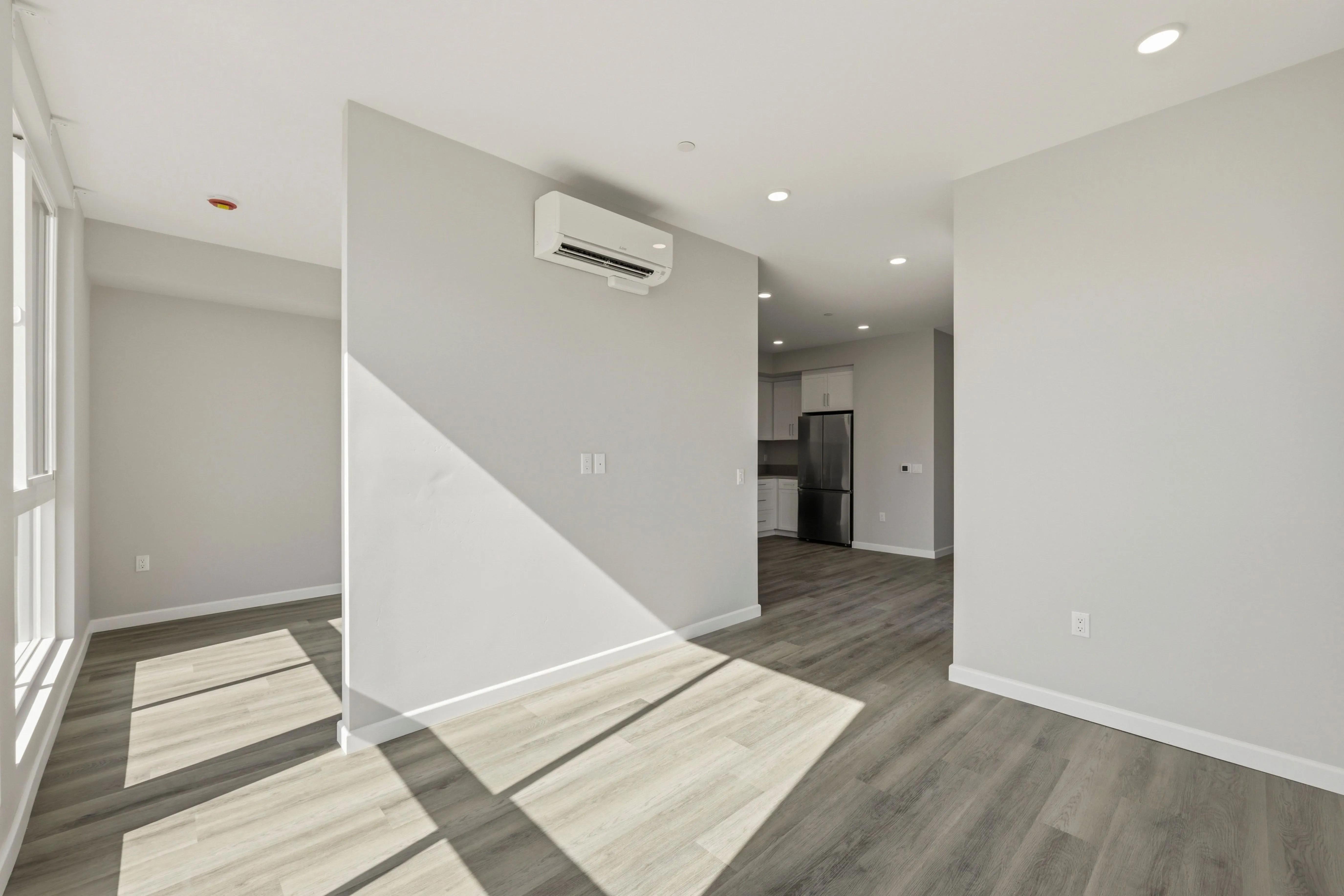 Empty modern apartment with grey wood floors, white walls, and kitchen with stainless steel fridge.