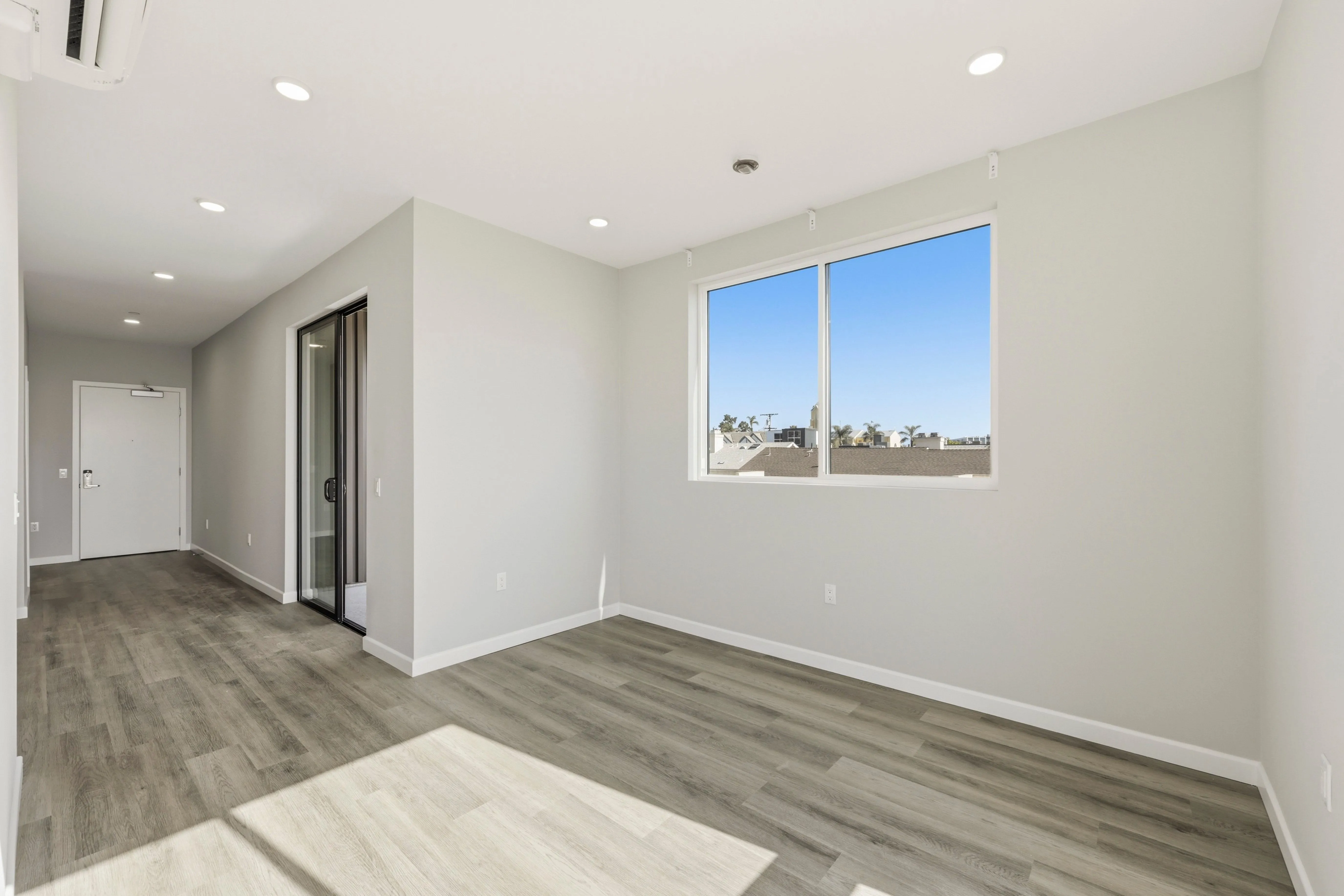 Empty room with wood floor, white walls, a large window, and hallway leading to a closed door.