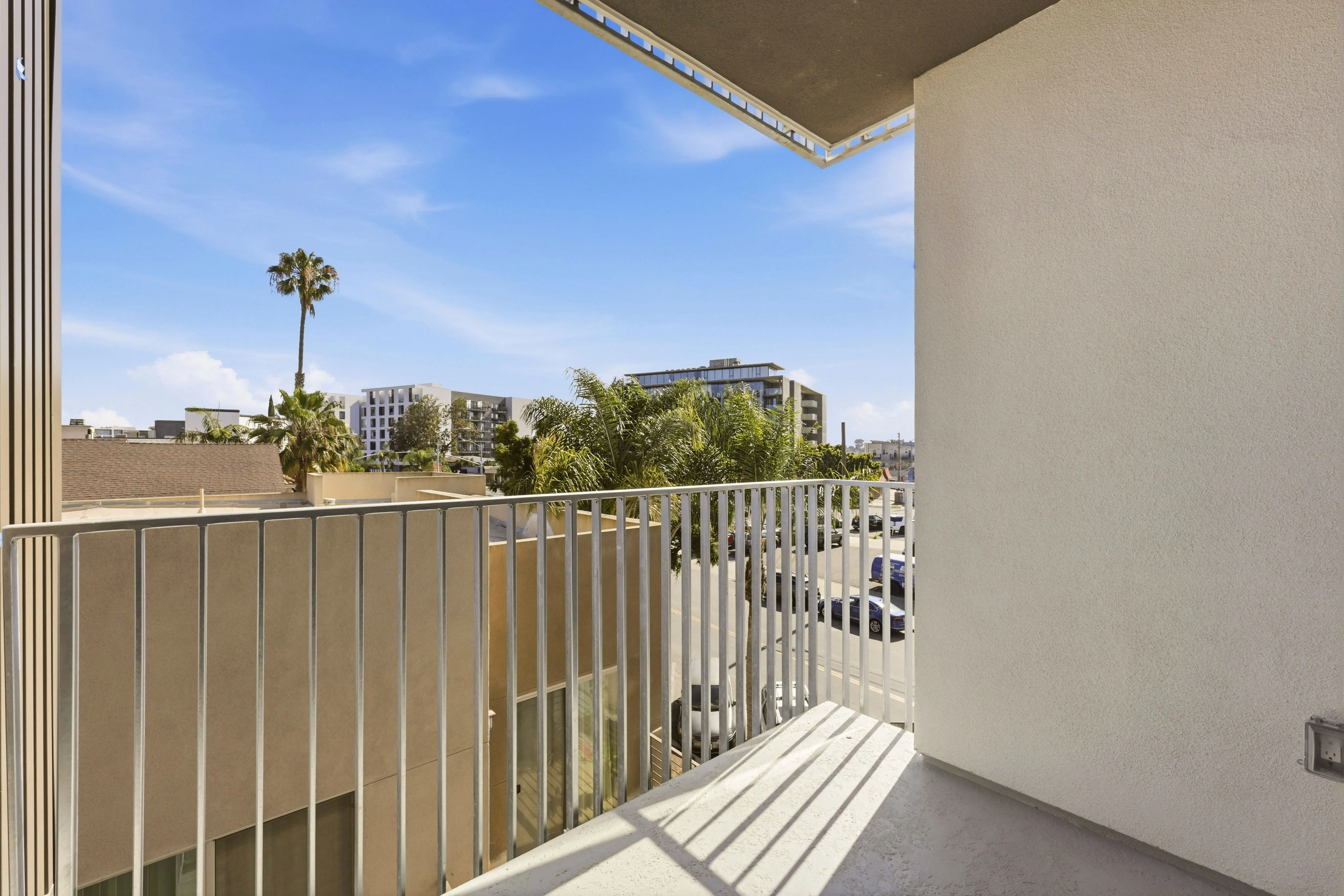 Small balcony with metal railing overlooking palm trees, parked cars, and modern buildings under blue sky.