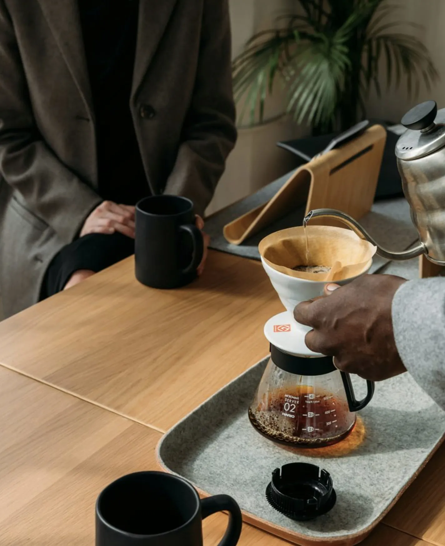Person pouring hot water from kettle into coffee dripper with filter on glass server at table.