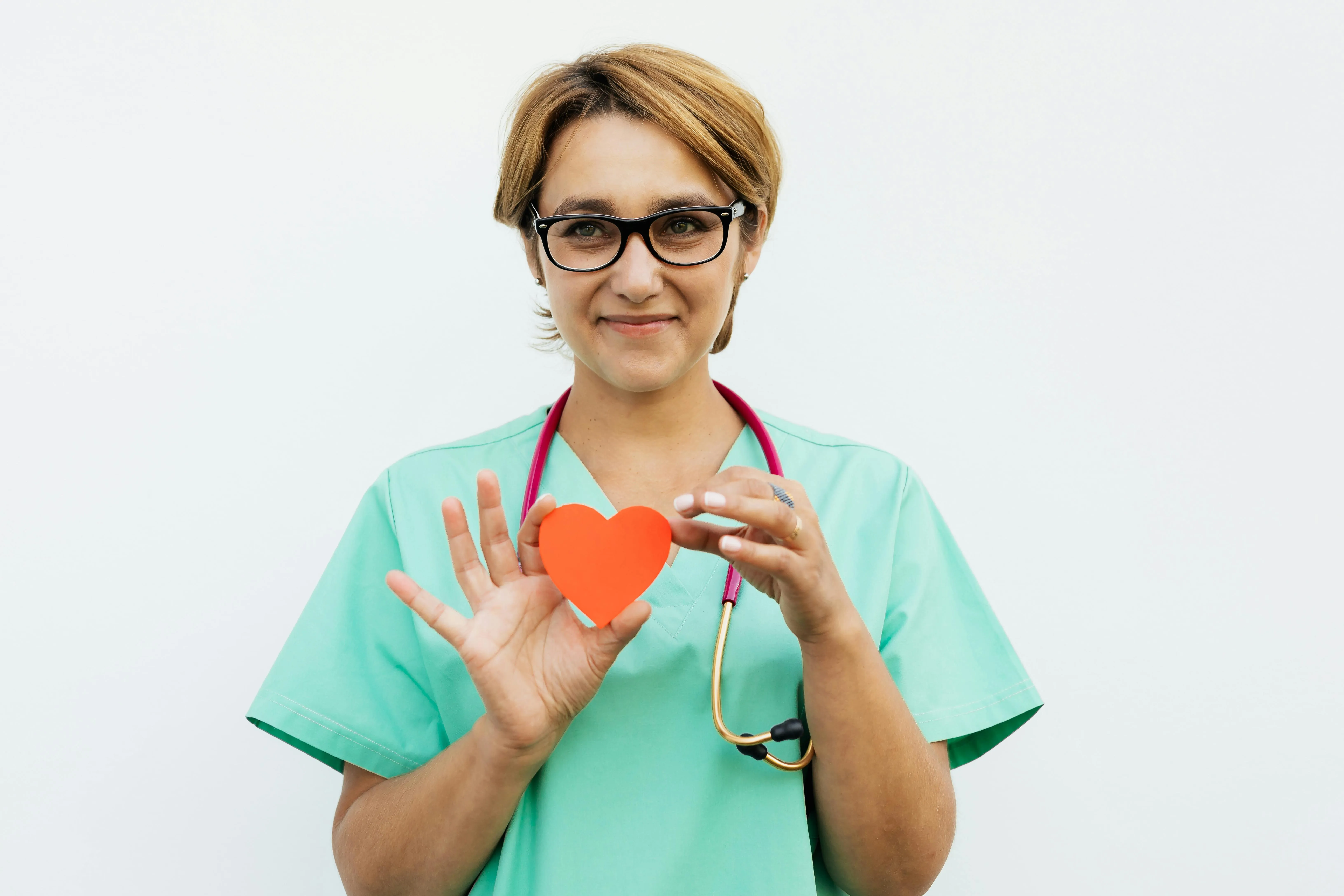Healthcare worker in scrubs with stethoscope holds a red paper heart symbolizing care.