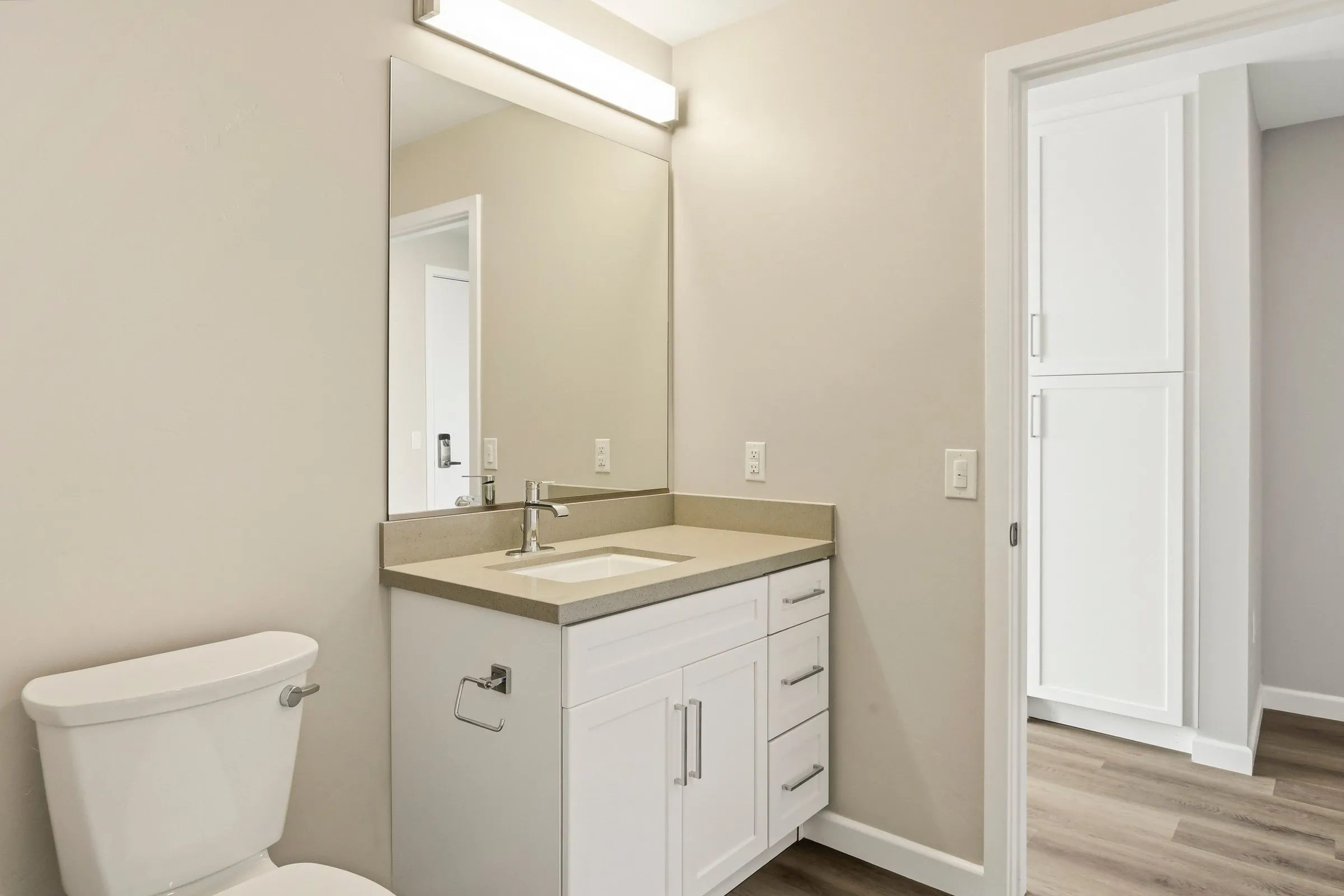 Modern bathroom with white vanity, rectangular sink, large mirror, toilet, and beige walls.