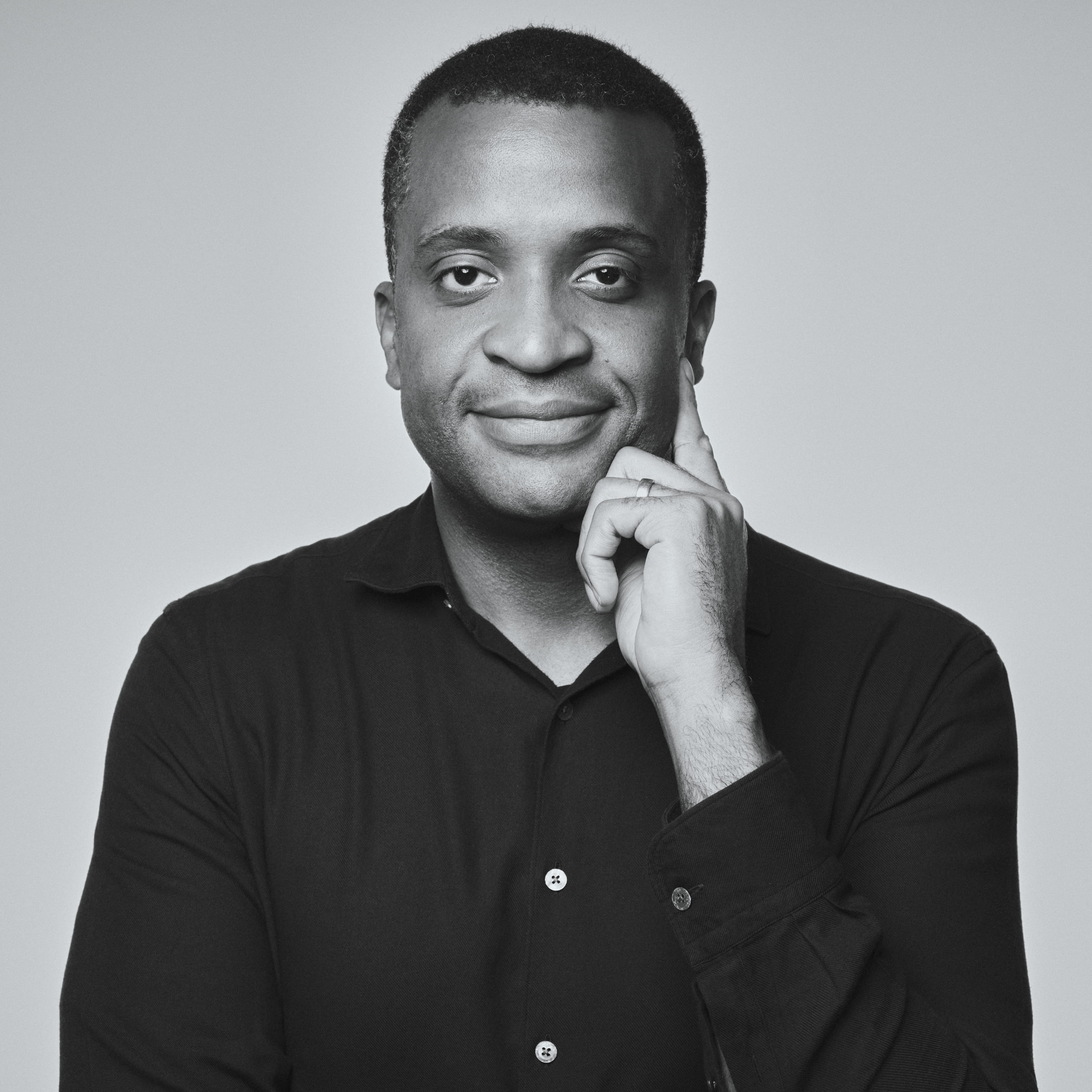 Founder, Obi Asika in a black shirt with finger resting on his cheek, smiling slightly against a plain background.