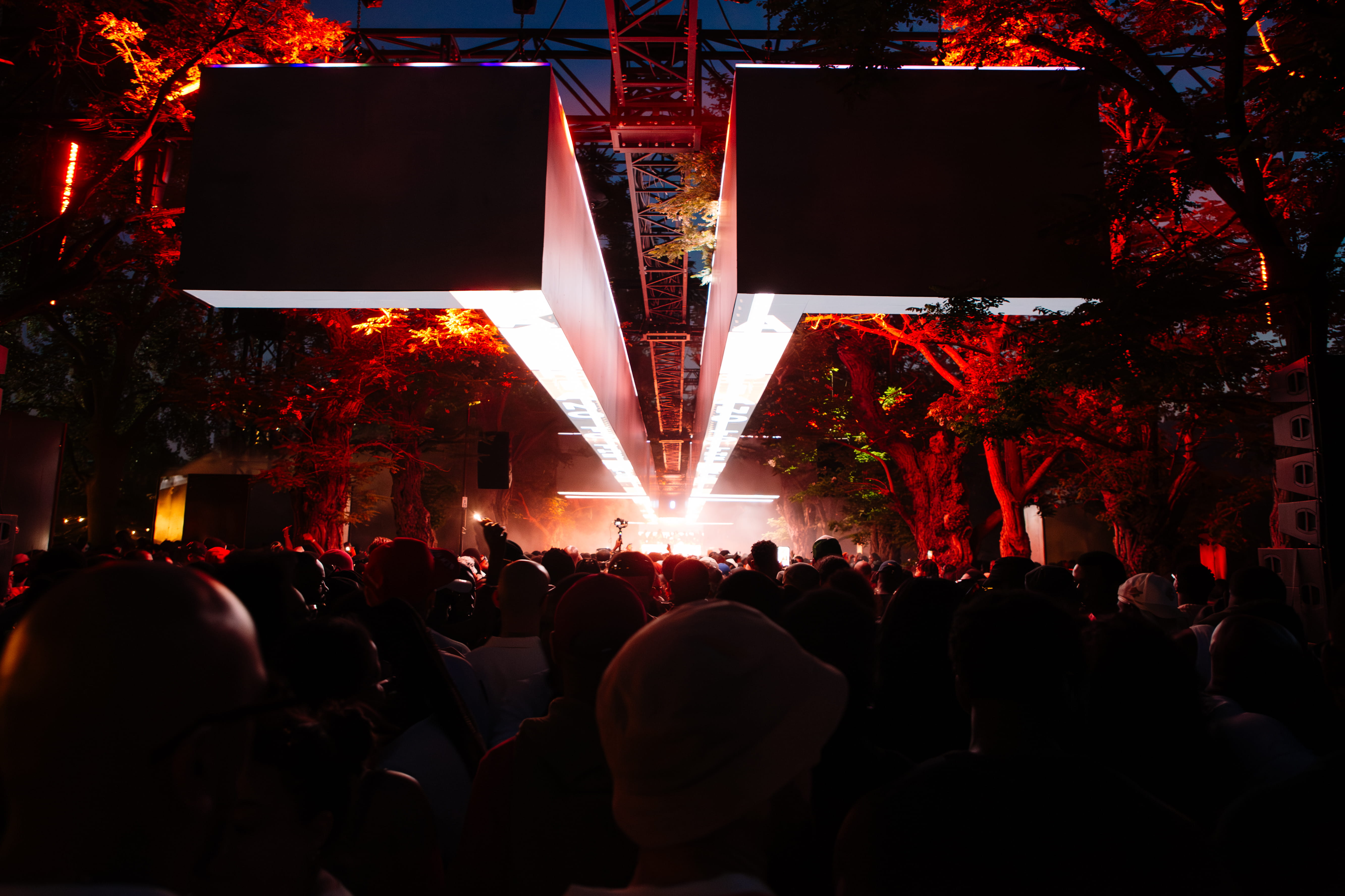 Crowd at night under red-lit trees and large suspended rectangular LED lights at an outdoor event.