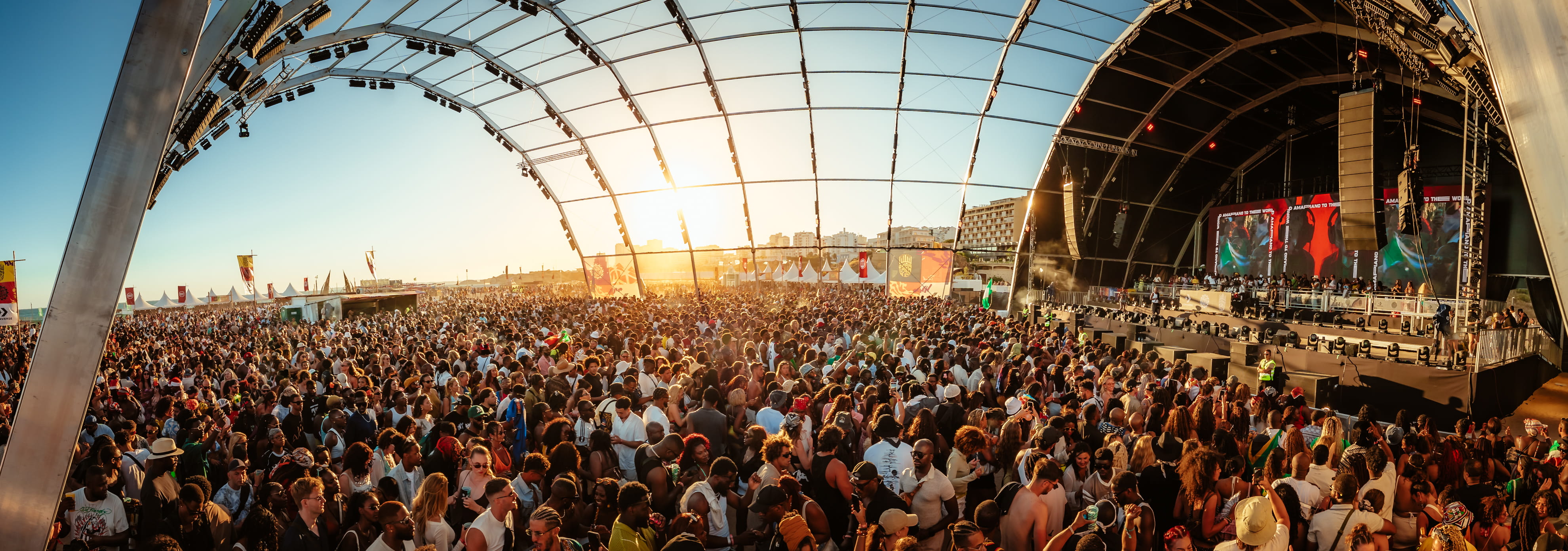 Large crowd enjoying a sunny outdoor music festival under a massive transparent stage canopy.
