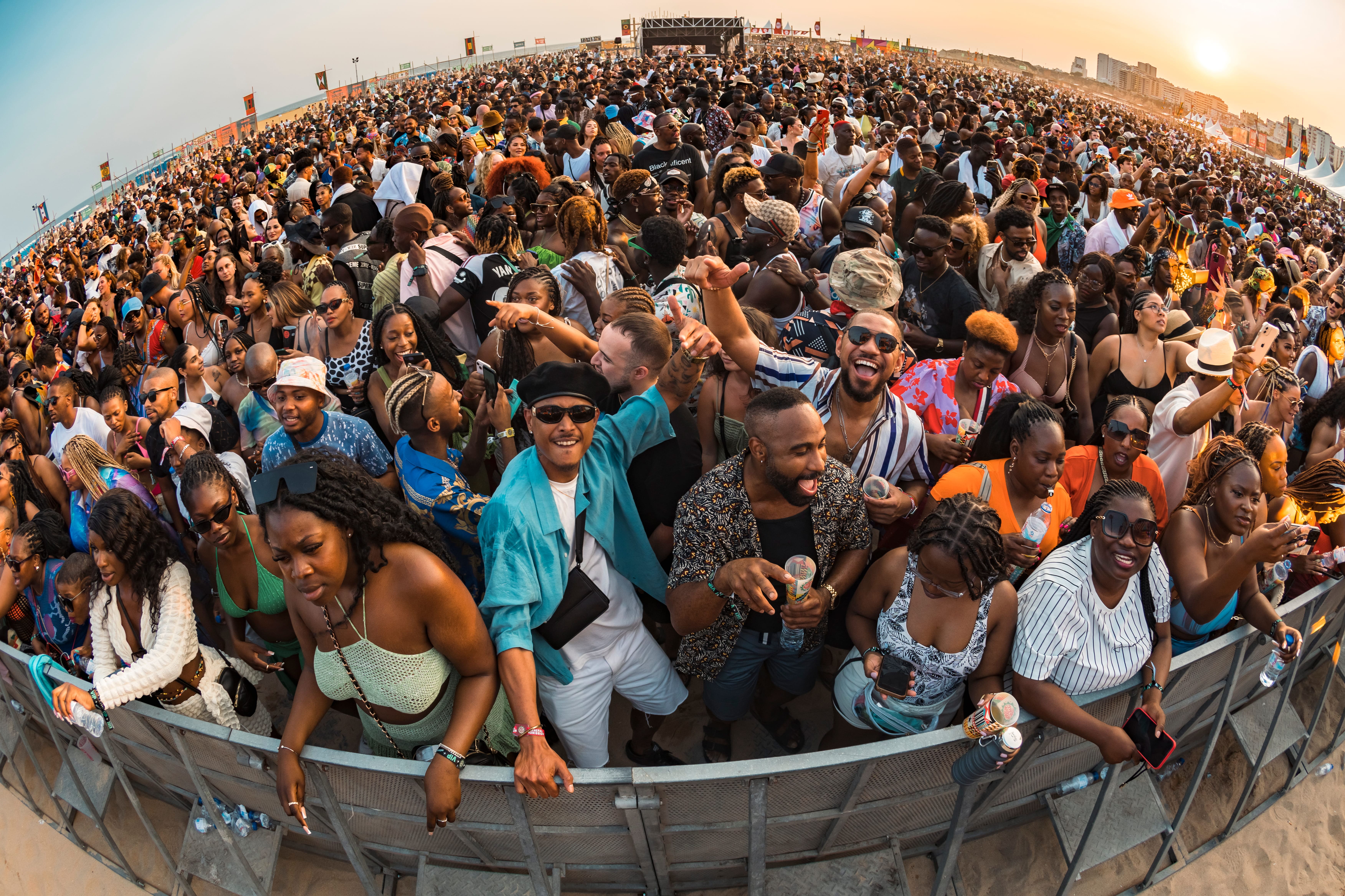 Crowded outdoor music festival with diverse people enjoying, dancing, and socializing at sunset.