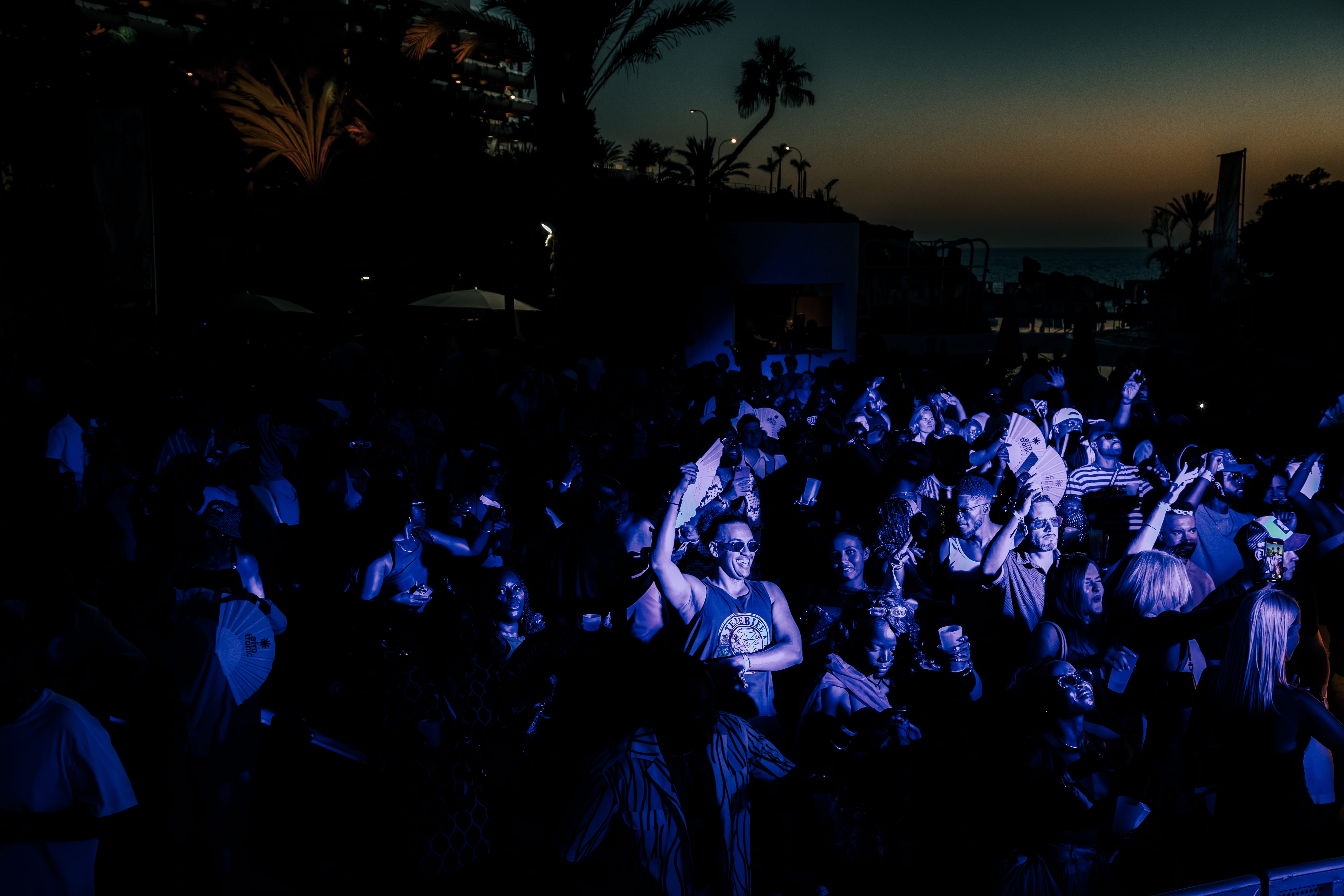 Crowd dancing and raising hands under colorful lights and smoke at a lively nighttime event.