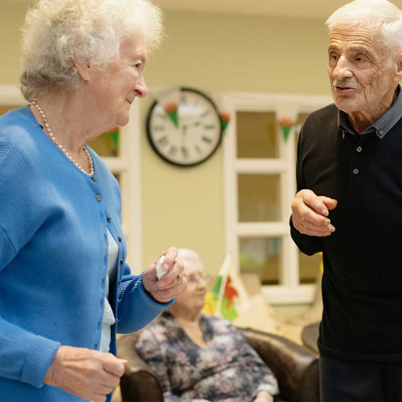 Elderly man and woman dancing and smiling indoors with another seated woman in the background.