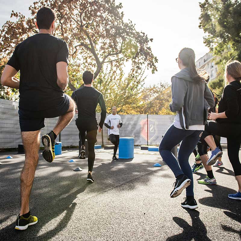 Group of people exercising outdoors, lifting knees while following an instructor against a sunny backdrop.