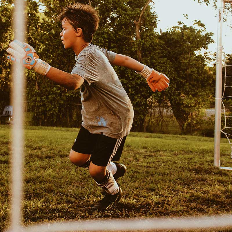 Young soccer goalkeeper wearing gloves and gray shirt poised to catch a ball on grassy field.