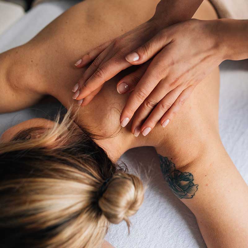 Close-up of hands massaging the upper back and neck of a woman with a floral tattoo.