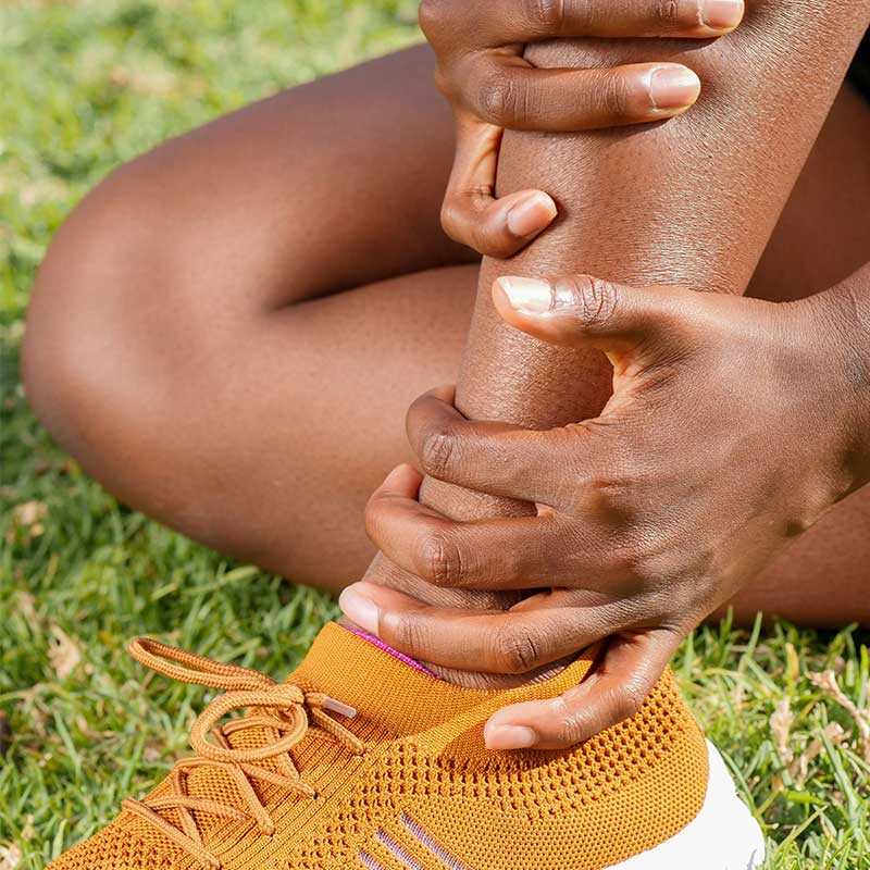 Person sitting on grass holding their lower leg and ankle, wearing orange athletic shoe, indicating leg or ankle pain.