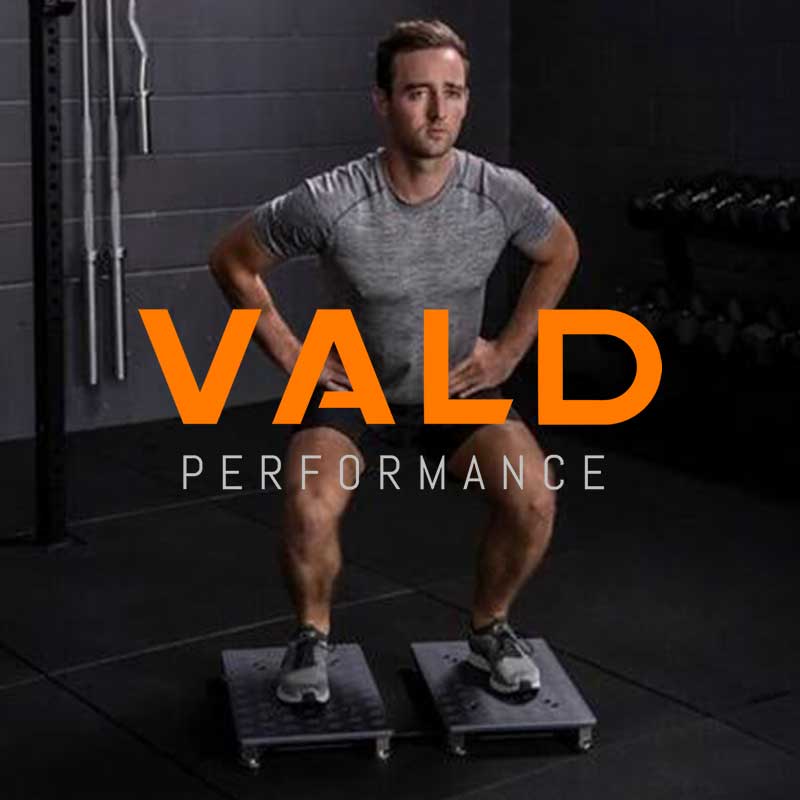 Man in gray athletic shirt and black shorts performing a squat exercise on two black platforms in a gym, with weights in the background and the text 'VALD PERFORMANCE' overlaid in orange and white.