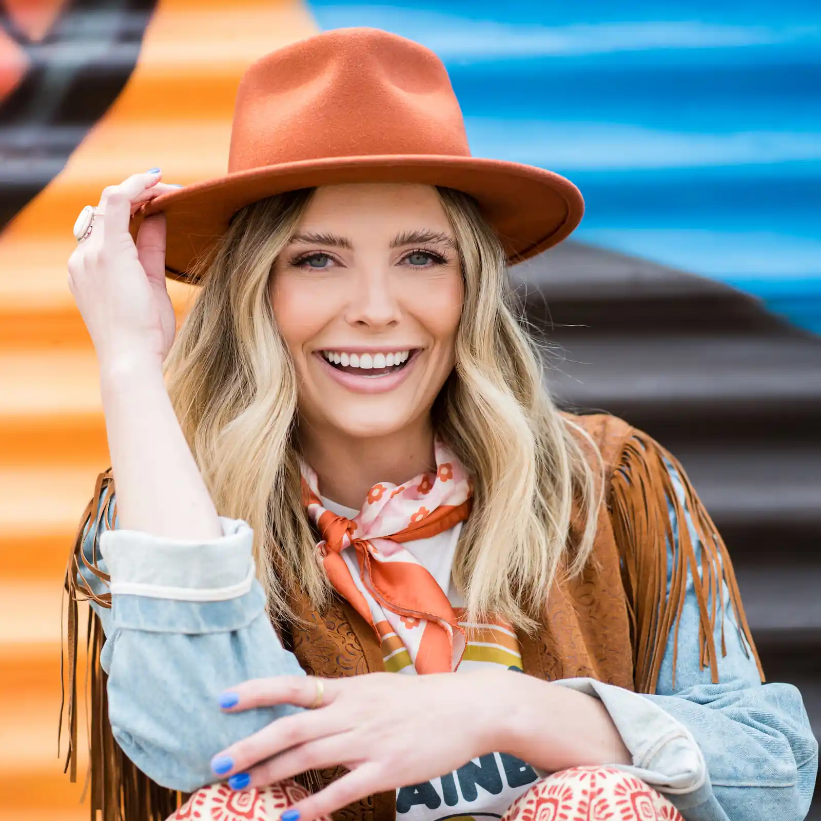 Smiling woman wearing a brown hat, fringed jacket, patterned scarf, and blue nail polish against a colorful striped background.