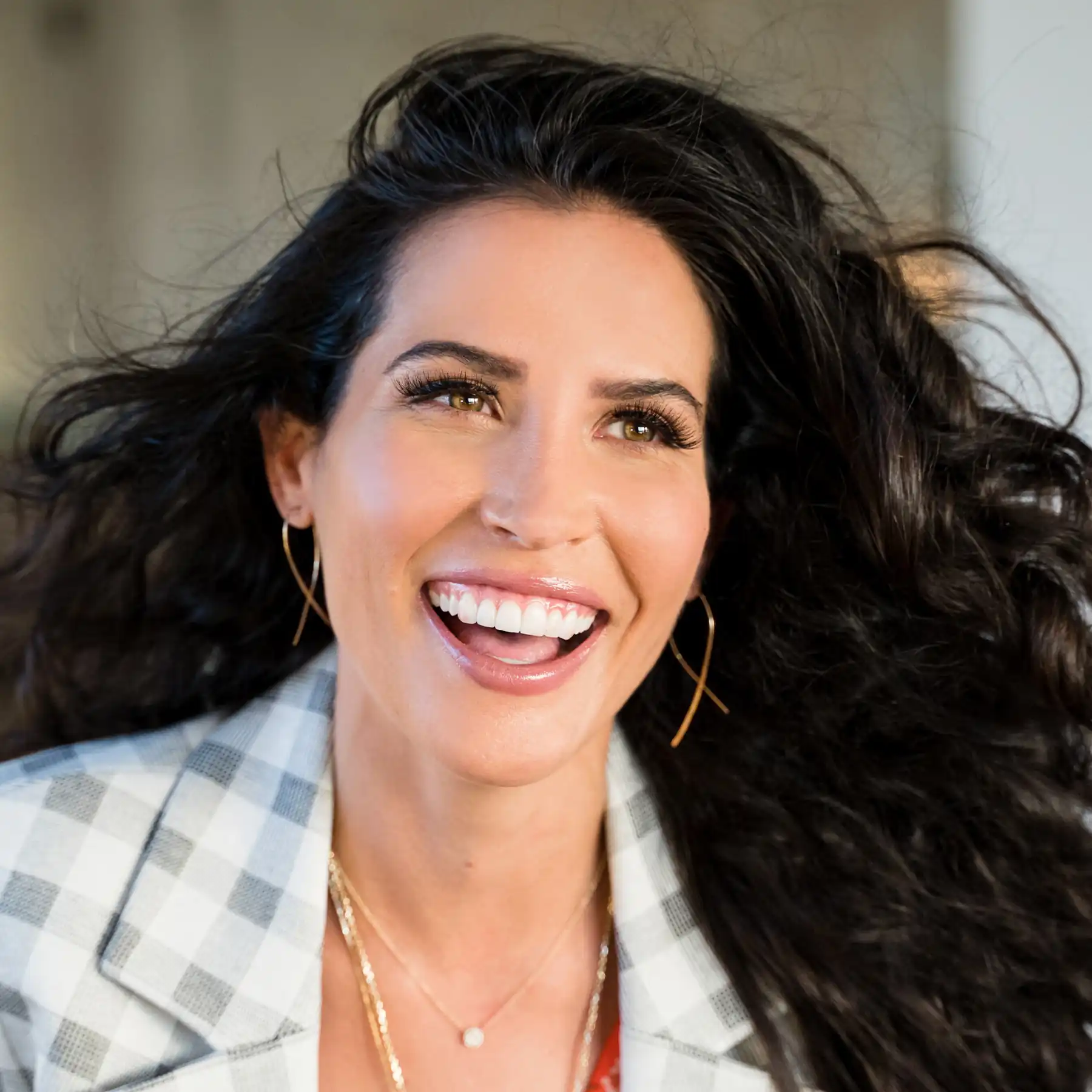 Smiling woman with long dark hair, wearing hoop earrings and a checkered blazer.