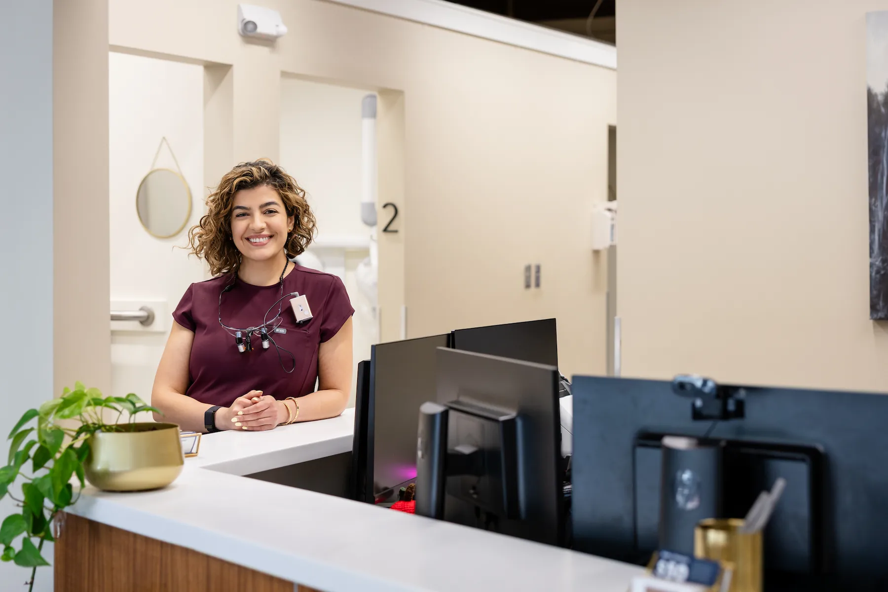 Smiling female dental staff standing behind reception desk with computer monitors and potted plant.