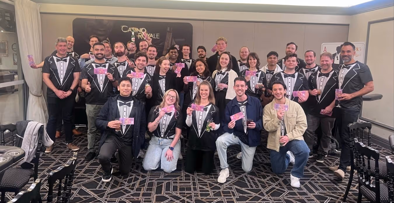 Group of people wearing tuxedo-themed shirts holding colorful notes and drinks in a room with a Casino Royale sign.