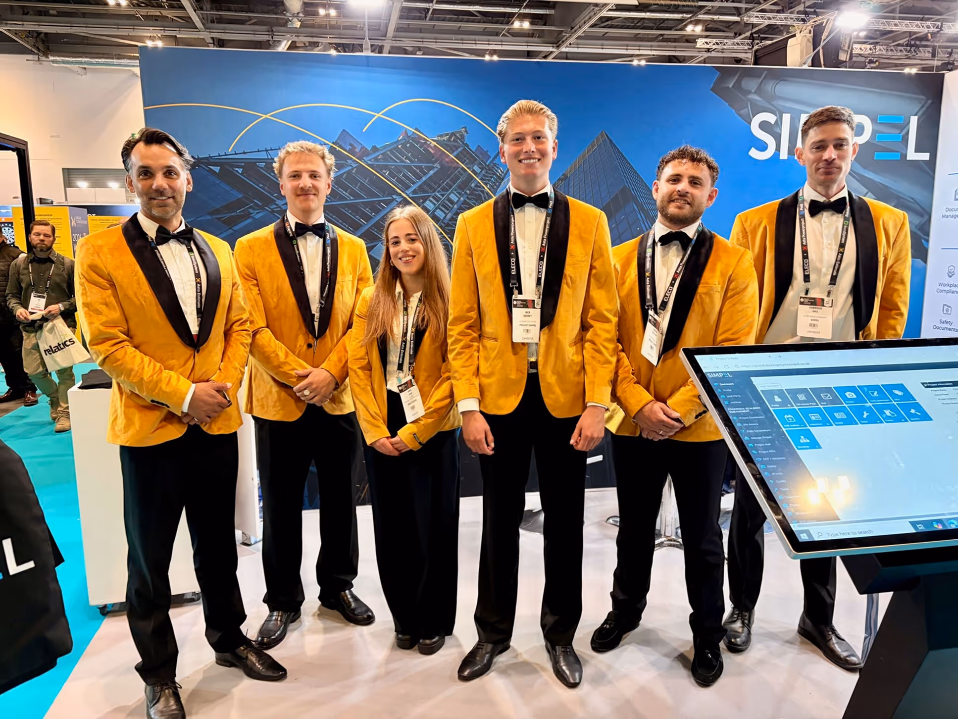 Six individuals in yellow blazers and black pants stand smiling at a trade show booth with a monitor.
