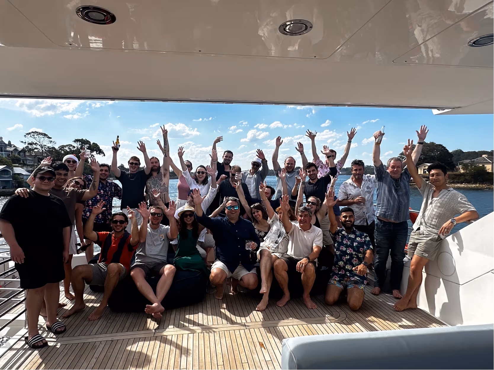 Group of people posing happily on a boat deck with hands raised, water and houses in the background.