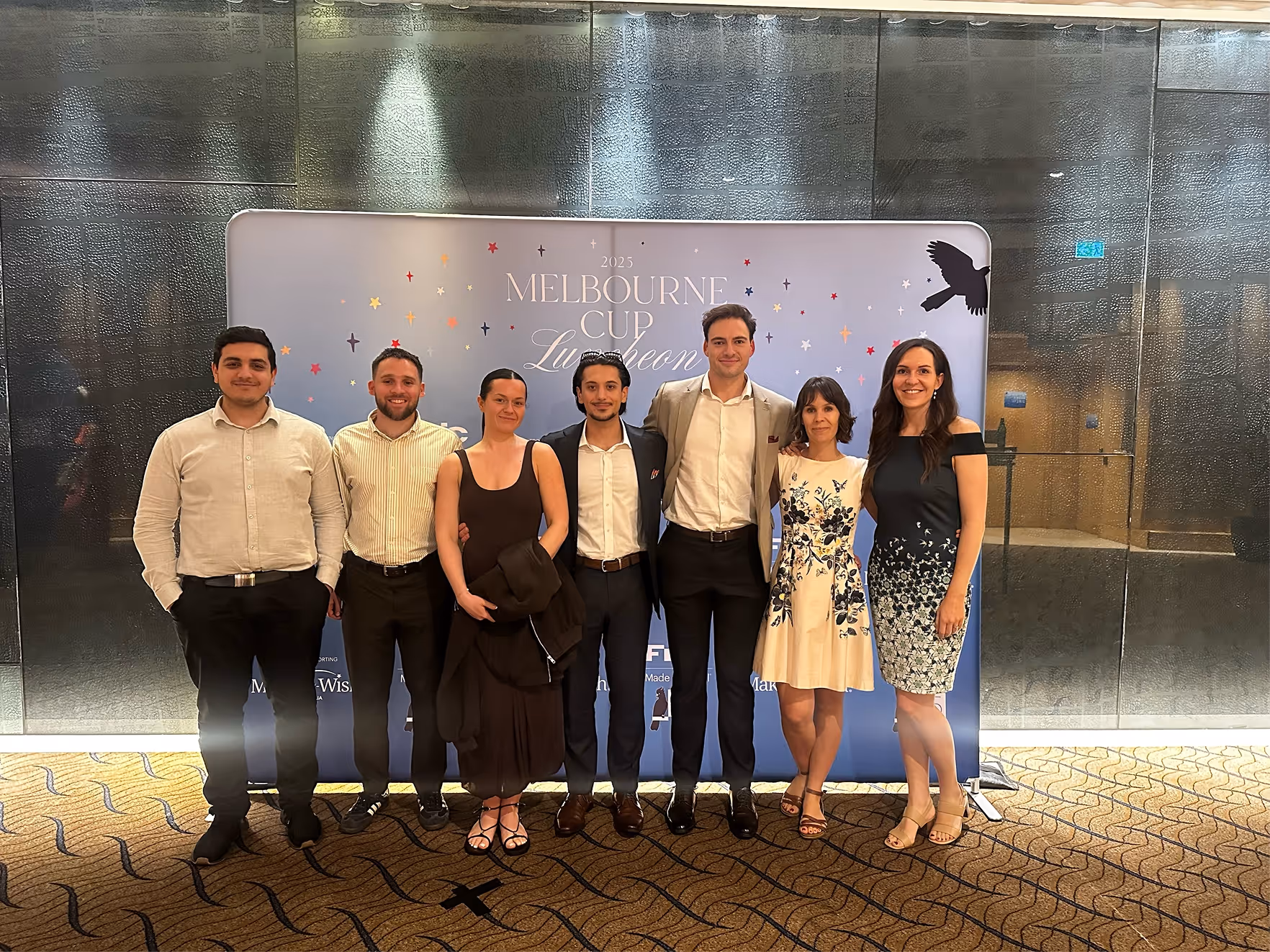 Seven people standing smiling in front of a 2025 Melbourne Cup Luncheon backdrop indoors.