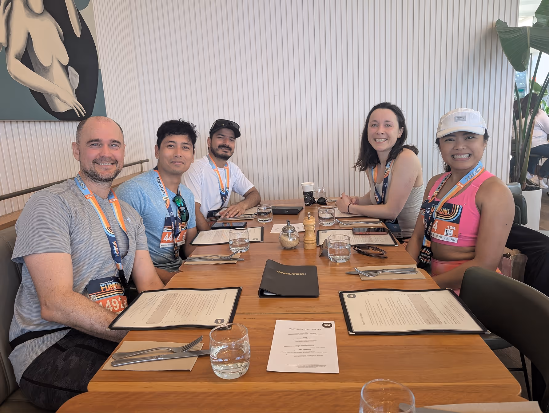 Five smiling people seated around a restaurant table with menus and water glasses, wearing race medals.