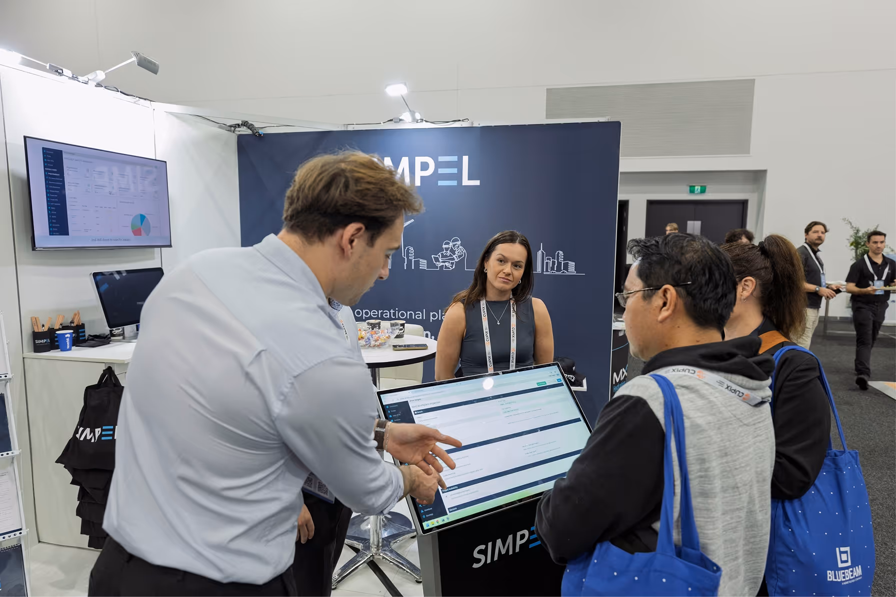 Man explaining software on a touchscreen at a SIMPEL booth to two conference attendees with Bluebeam bags.