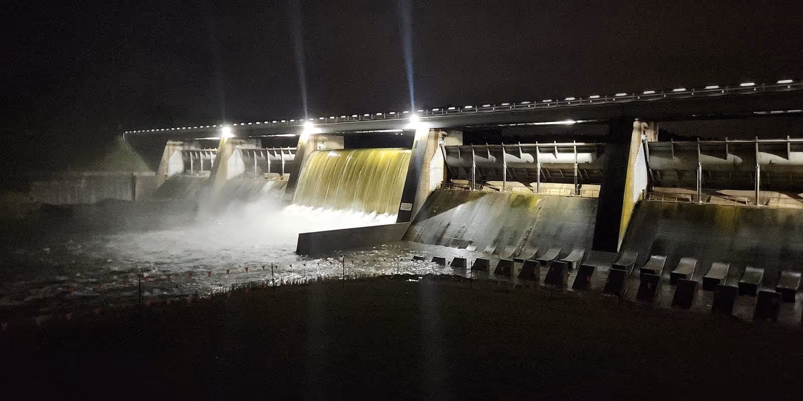 Night view of a dam spillway with water flowing over and bright lights illuminating the structure.