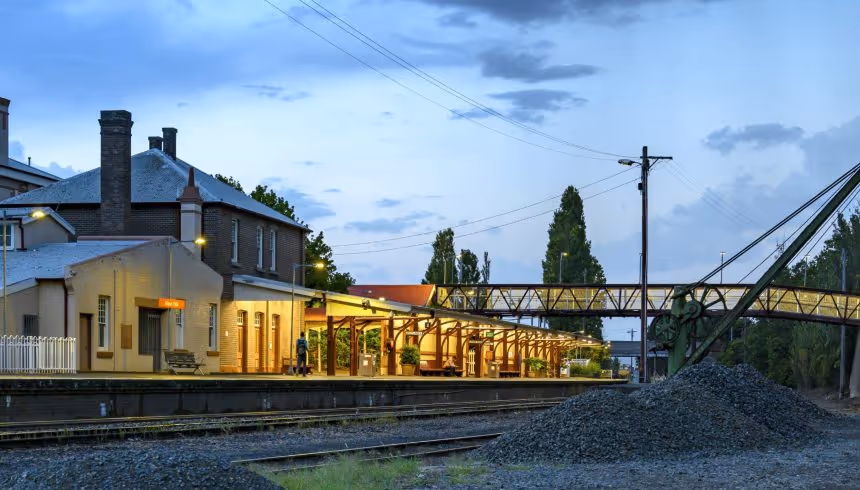 Evening view of a train station platform with lit canopy, brick buildings, train tracks, and overhead bridge.