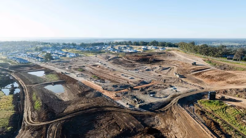 Aerial view of a large construction site with earthworks and residential housing in the distance.