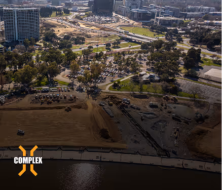 Aerial view of urban construction site near water with surrounding roads, buildings, and green trees.