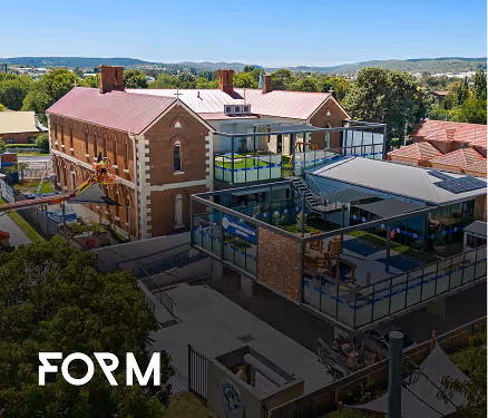 Aerial view of a brick building with modern glass extensions in a green suburban area under a clear sky.