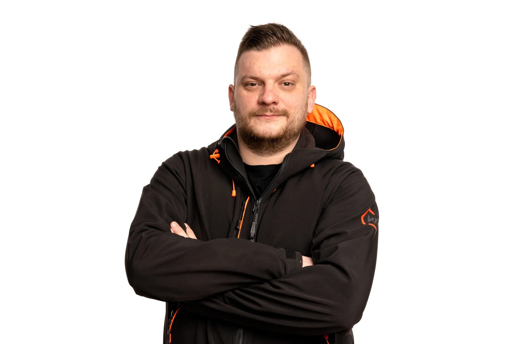 Mies mustassa takissa, jossa on oransseja yksityiskohtia, seisoo kädet ristissä valkoista taustaa vasten. Hänellä on parta ja lyhyet hiukset, ja hän katsoo kameraan hymyillen. Kuva on otettu tasaisessa studiovalaistuksessa.