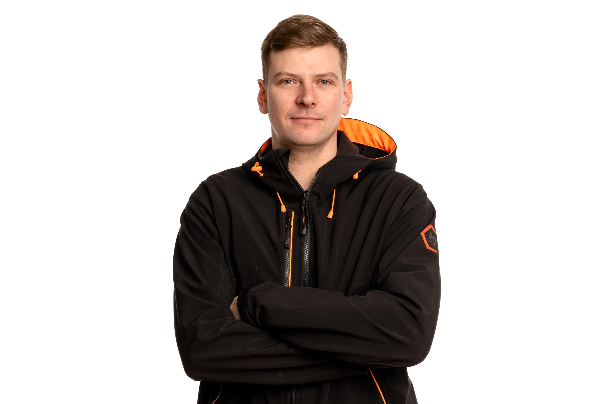 Mies seisoo valkoista taustaa vasten, katsoo kameraan. Hänellä on lyhyet ruskeat hiukset ja siniset silmät. Yllään musta takki oranssilla hupun vuorella ja oransseilla vetoketjun yksityiskohdilla. Kädet ovat ristissä rinnan päällä. Vasemmassa hihassa on oranssi kuusikulmainen logo.