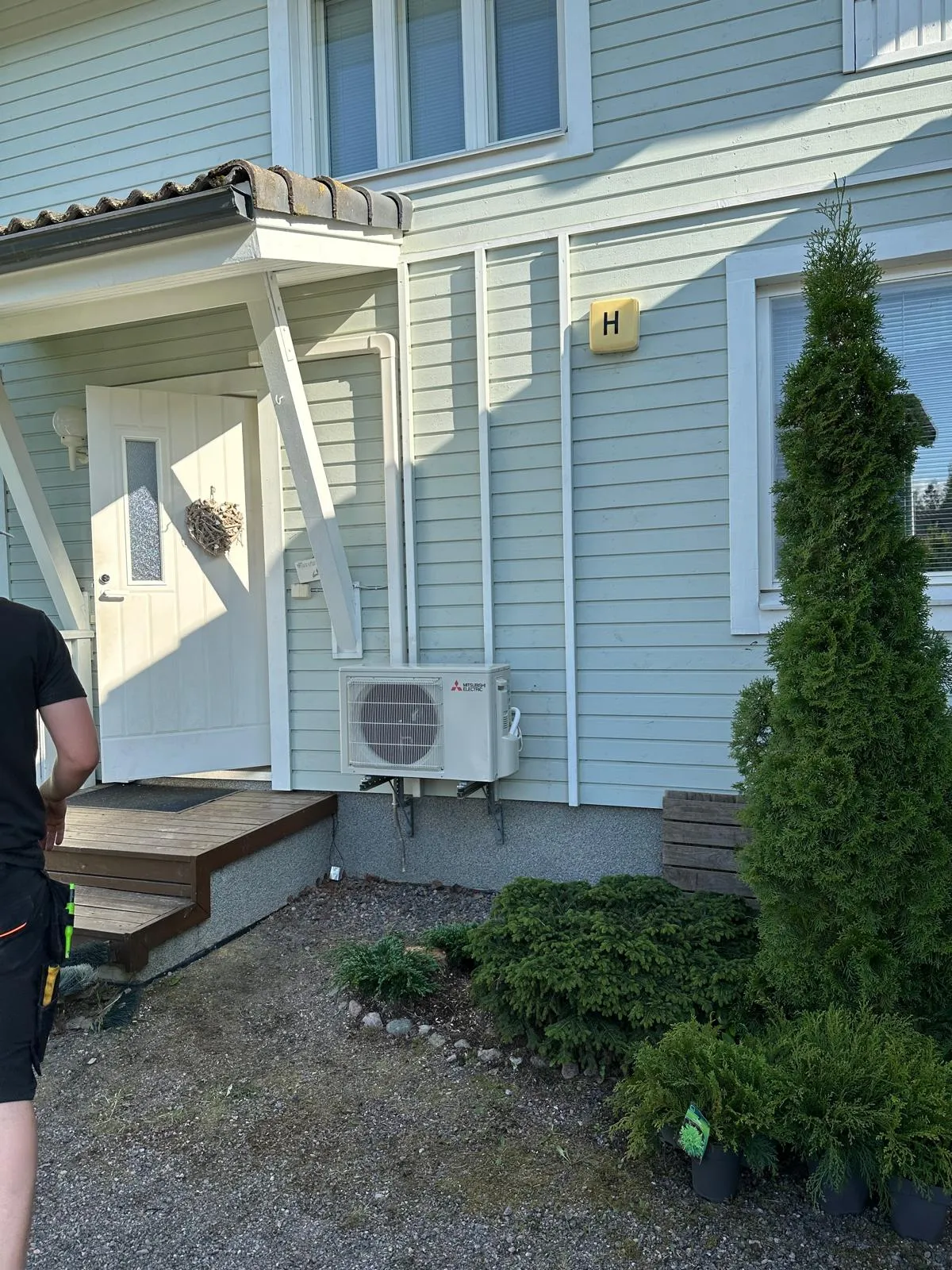 Vaaleansinivihreän talon ulkoseinä, jossa on valkoinen ovi, puinen kuisti ja Mitsubishi Electric -ilmalämpöpumpun ulkoyksikkö. Seinällä on keltainen H-kirjaimella varustettu kyltti. Talon edessä on vihreitä pensaita ja ruukuissa olevia kasveja sorapihalla. Kuvan vasemmassa reunassa näkyy osittain henkilö. Aurinkoinen päivä.