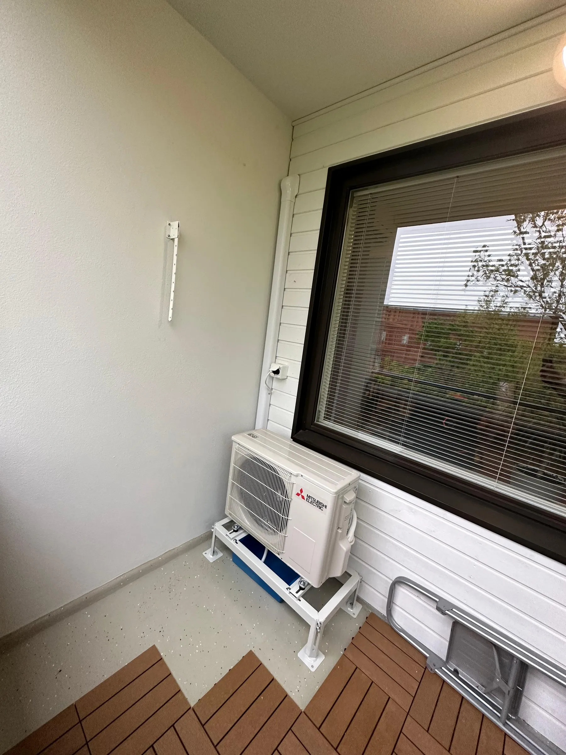 Valkoinen Mitsubishi Electric -ilmastointilaitteen ulkoyksikkö seisoo valkoisella metallitelineellä parvekkeella. Telineen alla on sininen astia. Vasemmalla on vaaleanharmaa rosoinen seinä ja oikealla valkoinen vaakapaneeliseinä tummanruskeakehyksisen ikkunan ympärillä, jossa on sälekaihtimet. Seinien nurkassa kulkee valkoinen putki, ja sen vieressä on pistorasia, johon on kytketty johto. Lattialla on vaaleanharmaa pilkullinen pinnoite, osittain peitettynä ruskeilla, toisiinsa lukittuvilla laatoilla. Vasemmalla seinällä on valkoinen metallikiinnike ja oikealla lattialla taitettu metallinen jakkara. Kuva on otettu kirkkaassa päivänvalossa.